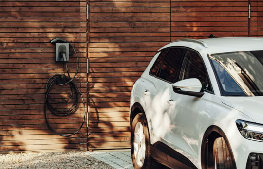 Silbernes Auto vor einer Holzwand mit einer Ladestation und einem aufgerollten Kabel.